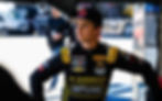 Young race car driver in a black suit with sponsor logos, stands confidently in a pit area. Background shows blurred vehicle and person.