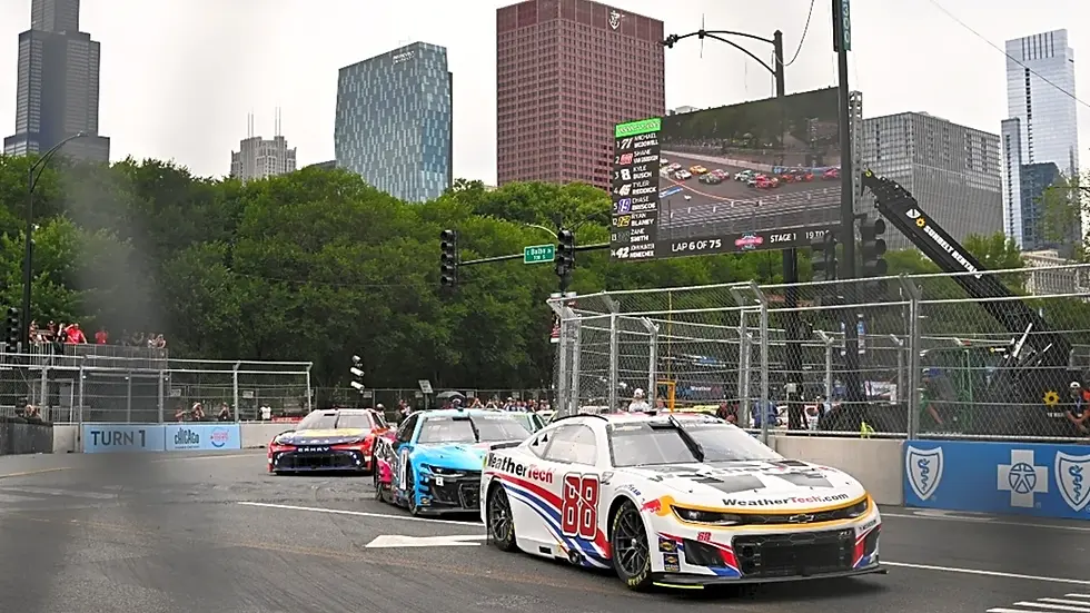 Race cars on a track in a city setting, with a digital leaderboard in the background showing rankings. Sky is overcast; cars have vivid colors.