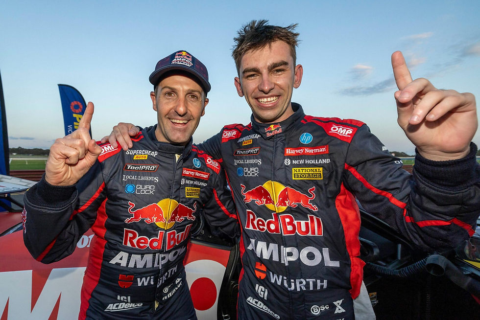 Two race car drivers in Red Bull Ampol suits smile, holding up index fingers, in front of a car at a racetrack.