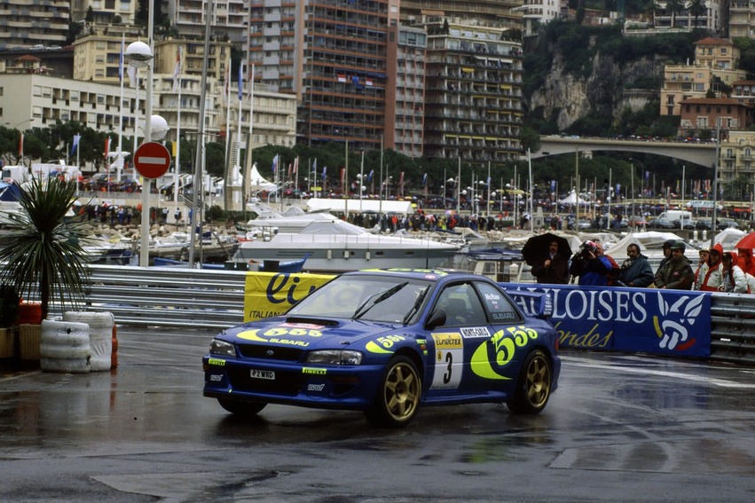 Blue rally car with yellow decals skids on wet street in cityscape, crowds watching under umbrellas, waterfront and buildings behind.