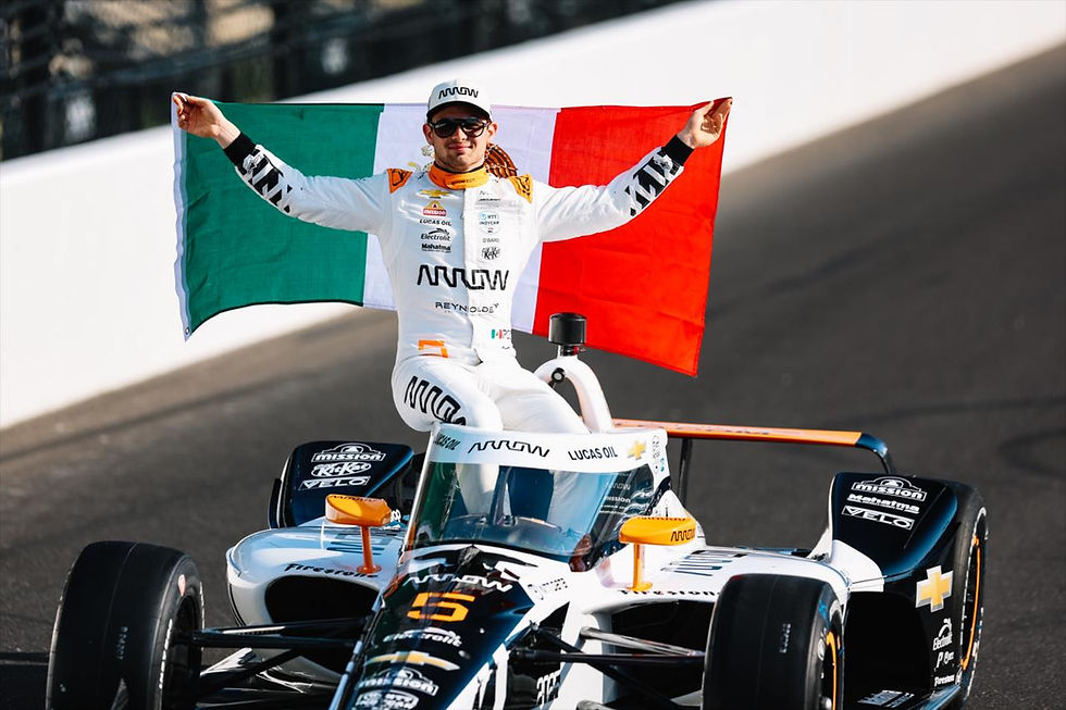 Racer in white suit holds Mexican flag, stands on IndyCar with "ARROW" and "LUCAS OIL" logos. Bright track setting, triumphant mood.