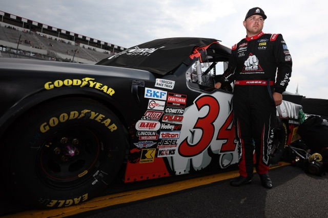 Layne Riggs stands beside a black car numbered 34 at a Pocono Raceway. Cloudy sky and grandstands visible.