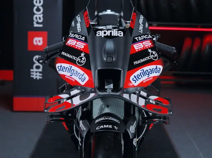 Front view of a racing motorcycle with prominent sponsor logos like Aprilia and Sterilgarda. The bike is black and red, set indoors.