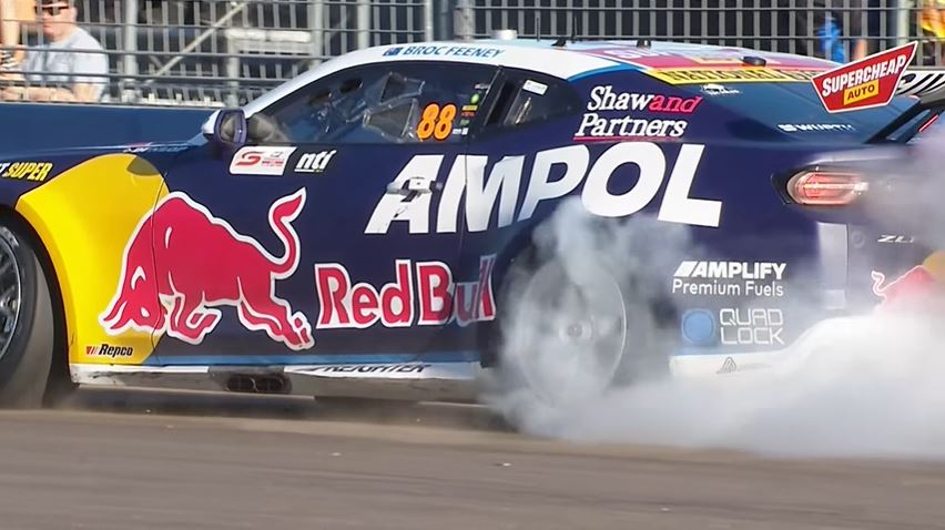 Racing car with Red Bull and Ampol logos doing a burnout, smoke billowing from rear tire. Track setting, vibrant blue and yellow design.