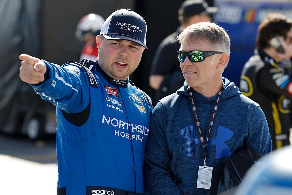 Race car driver in blue suit and cap points ahead while talking to a man in sunglasses and hoodie. "Northside Hospital" visible.