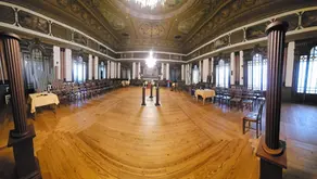 Grande salão interior com piso de madeira, cadeiras alinhadas ao longo das paredes, janelas altas em arco e tecto decorado; ao centro vê-se um pequeno estrado.