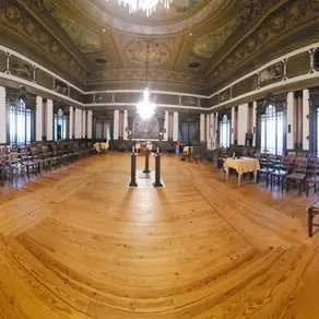 Grande salão interior com piso de madeira, cadeiras alinhadas ao longo das paredes, janelas altas em arco e tecto decorado; ao centro vê-se um pequeno estrado.