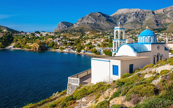 A picturesque whitewashed Greek church with a blue dome sits on a hillside overlooking a calm bay and a charming village nestled against a backdrop of mountains. The scene is bathed in bright sunlight, showcasing the vibrant colors of the buildings and the clear blue water.