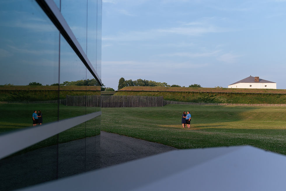 Fort Wellington in Prescott, Ontario at sunset - we see a couple standing in a ray of light in the distance. Their reflection on the side of the welcome centre, with the fort in the background.