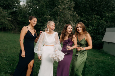 Braut steht lachend mit ihren Freundinnen in farbenfrohen Kleidern auf einer Wiese.