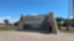 Person posing by Big Cypress National Preserve sign, with a clear blue sky and palm trees in the background. The mood is cheerful.