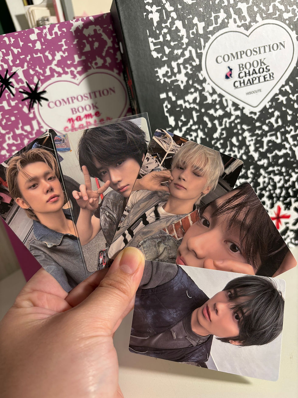 A close-up photograph of a hand holding several TOMORROW X TOGETHER (TXT) photocards in a fan-shaped arrangement, with "The Chaos Chapter: FREEZE" album books visible in the background.