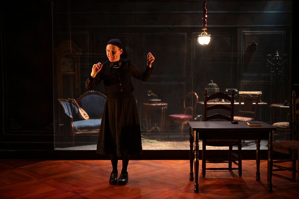 An actor wearing a black dress stands on a set filled with antique furniture, a mischievous expression on their face