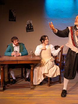 Three people sit at a table, watching a man standing with his arm outstretched, seemingly in the middle of a dramatic monologue