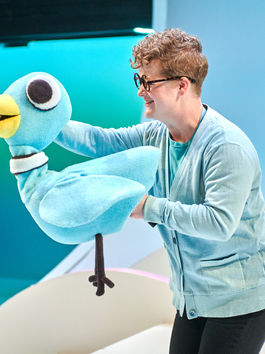 An actor wearing glasses controls a large, cartoonish, blue bird puppet