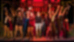A man stands center stage in a plain costume, while around him actors in bright, colorful costumes strike a jazzy pose in front of a red background
