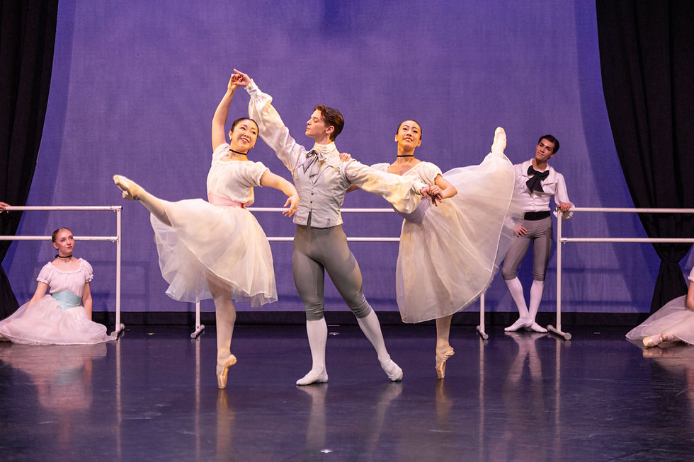 A male dancer stands center, holding up two female dancers on pointe on either side of him, while other dancers look on in a ballet studio
