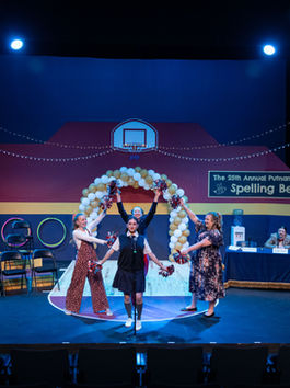 In a gym decorated with balloons, four women stand with their arms out in poses, holding pompoms, while three men look on from the sidelines