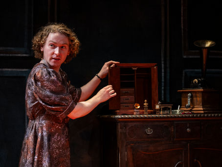 A person in a dress stands next to a dresser, upon which are miniature furniture items