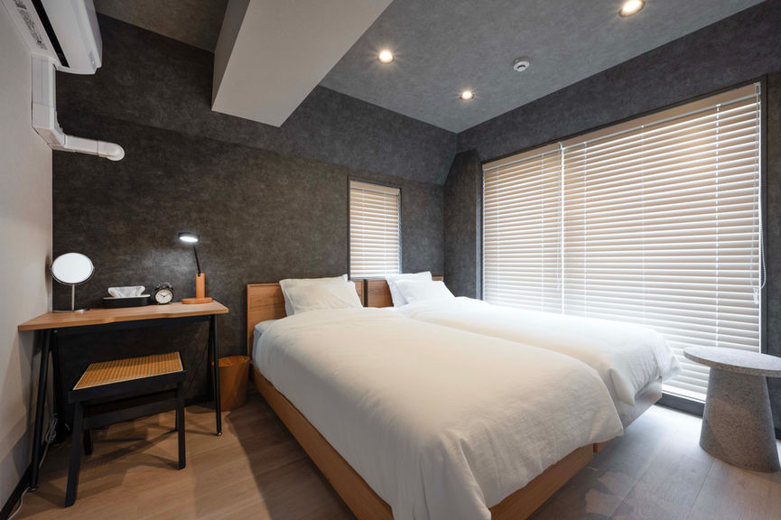 Room 1 twin bedroom with balcony access, two single beds in white linen, wood-tone desk, and air conditioning in a modern gray and white interior