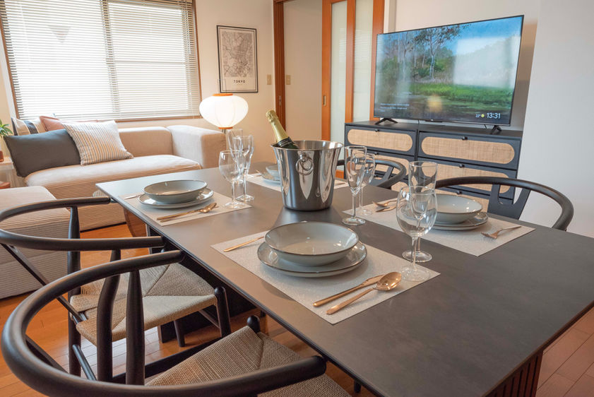 Dining table set with plates, cutlery, wine glasses, and wine bottle, with 55-inch TV and fabric sofa visible in the background