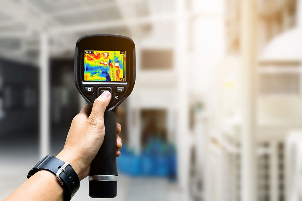 Hand holding a thermal camera showing a colorful heat map display in an industrial setting. The background is blurred and bright.