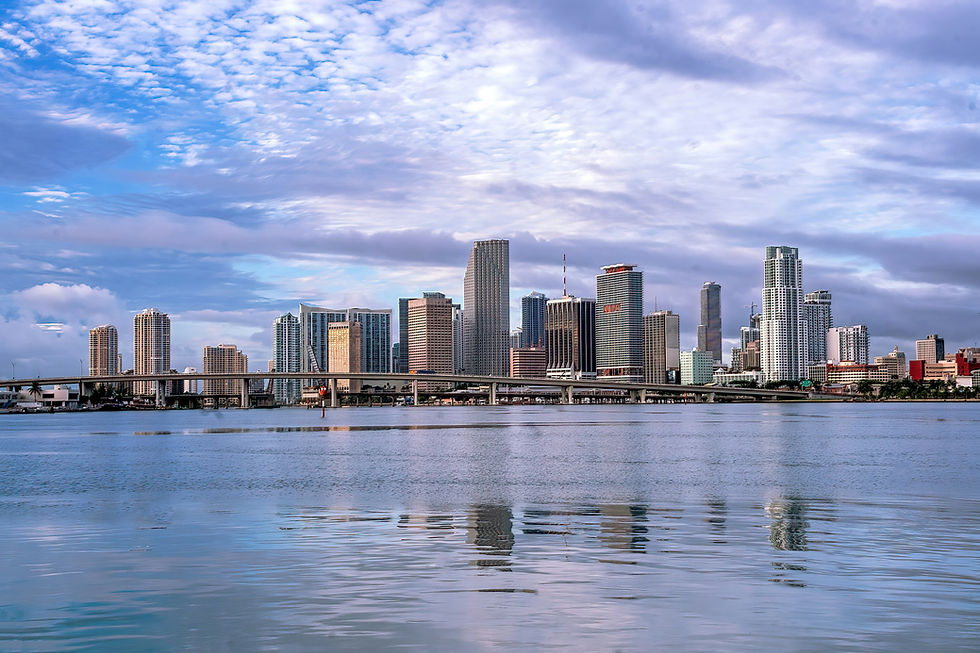 Skyline of modern city with tall buildings under a partly cloudy sky, reflected in calm waterfront. Bridge in foreground, serene atmosphere.