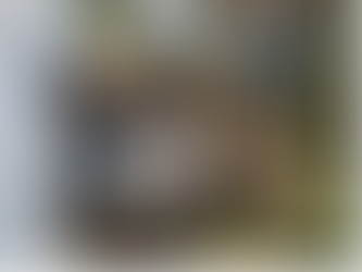 Thermal camera displaying a colorful heat map, held by a person in a workshop. Blur of people working in the background.