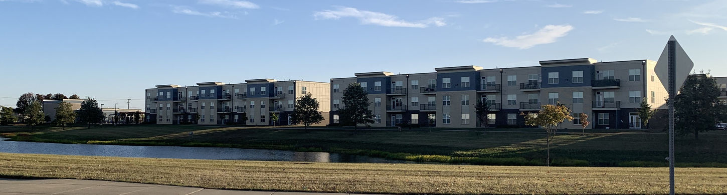 Century Apartment complex on the other side of a man-made pond and green grass. Above the building is a clear, blue sky.