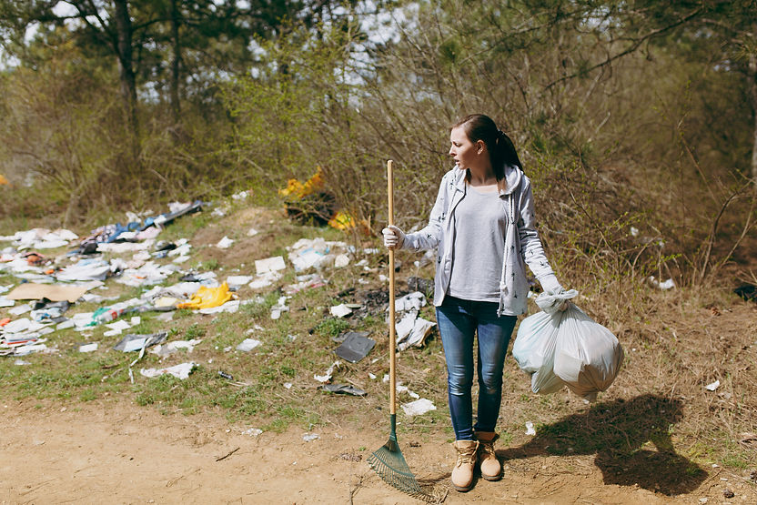 Young upset shocked woman cleaning holding trash bags and rake for garbage collection in littered pa