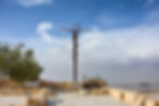 Sculpture of a twisted metal staff with a snake in a stone plaza, clear blue sky background, olive tree on the left, text on a sign.