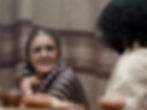 A woman in a striped shawl smiles warmly, sitting next to a person with curly hair. Brown striped curtain in the background. Cozy atmosphere.