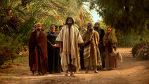 Group of robed people walk down a dirt path through palm trees, led by a bearded man in beige; solemn, historical scene.