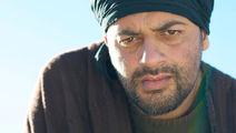Man with intense expression, wearing a black headscarf and dark clothing, against a clear blue sky background. Mood is serious.