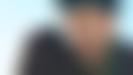 Man with intense expression, wearing a black headscarf and dark clothing, against a clear blue sky background. Mood is serious.