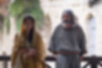 Two people in ancient attire stand by a wooden railing. The woman wears a yellow shawl, the man a grey robe. Stone walls with greenery in the background.