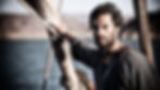 Bearded man on a sailboat gripping a rope, staring ahead with a serious look, sea and shore behind him.