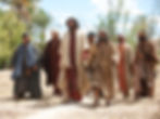 A group of men in robes walk on a dirt path surrounded by trees under a clear sky. They appear engaged and content. Earth tones dominate.