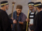 Four men in robes and hats, three holding staffs, gather closely. One examines his palm intently. The scene has an ancient, solemn feel.