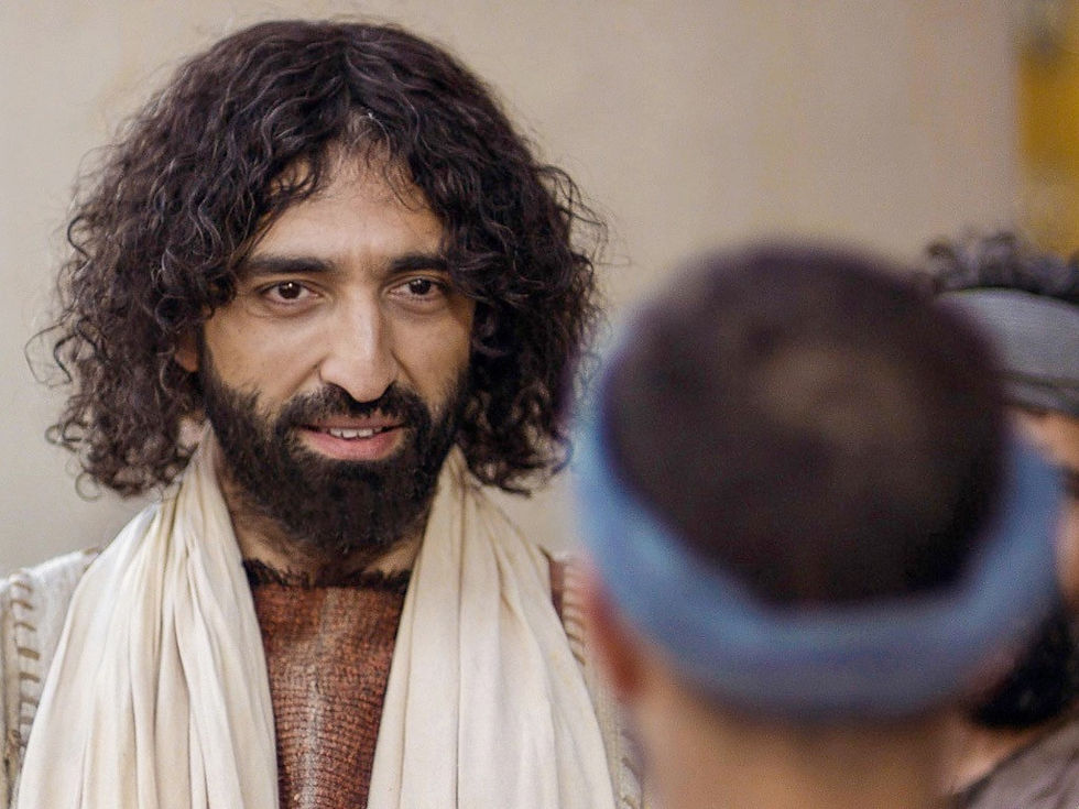 Man with curly hair and beard in a white scarf talks to another person. Neutral indoor background, soft lighting, focused expression.