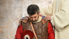 A man in Roman armor looks down, holding a helmet. A figure in white robe offers comfort, hand on his shoulder, against a textured wall.