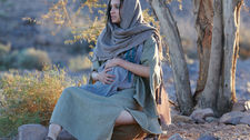 Woman in a hooded robe sits on rocky ground, hand on her belly, under a tree in a desert setting. Calm, contemplative mood, soft lighting.