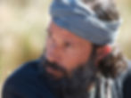 Man with a gray headscarf looking intently to the side. He has a beard and wears dark clothing. Bright, blurred background. Moody expression.