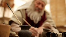 An elderly person in traditional attire writes with a quill at a wooden desk. Clay pots and scrolls are in the foreground, creating a historic setting.