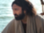 A man with long hair and a beard gazes thoughtfully out over water, wearing a beige cloak. The setting is calm and serene.