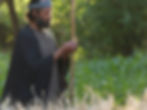 A man in a robe examines wheat stalks in a sunny, green field. He holds a walking stick and wears a headband, evoking a serene mood.