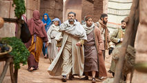 A group of people in historical clothing walk through a narrow, ancient stone street. Textiles hang in the background, creating a rustic scene.