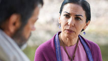 Serious woman in purple shawl and pendant talks with a blurred man outdoors, with soft natural background.