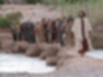 A group of men in robes walks across a rocky path by a river in a serene, natural setting, displaying a contemplative mood.