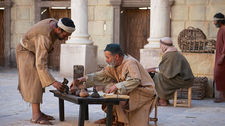 Men in robes exchange goods at a low table in an ancient stone courtyard. One man sits while another stands beside him. Rustic atmosphere.